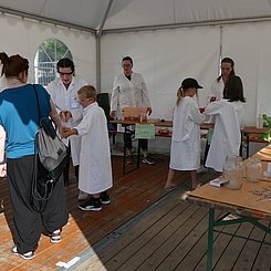 Blick in das Pagodenzelt des Lehr-Lern-Labors "Kinder experimentieren" bei EFFEKTE 2023 - auf Biertischen in einem U sind Experimente aufgebaut, dazwischen Kinder und Studierende in weißen Laborkitteln und mit Schutzbrillen.