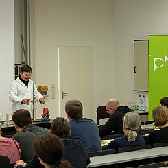 Ein dozent führt in einem Hörsaal am Pult ein Chemieexperiment für den Schulunterricht vor. Lehrkräfte sitzen im Hörsaal. Links ist ein grünes RoppUp mit PH Logo