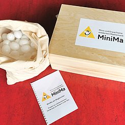 Geschlossene Materialkiste aus hellem Holz mit MiniMa Logo. Links daneben ein weißer Sack mit Plastikeiern.