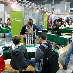 Auf den Science Days steht erklärt eine PH Studentin vor einem grünen PHKA RollUp mit Logo sitzenden Kindern mit einer Zahlentafel ein mathematisches Experiment