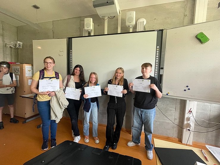 Fünf jugendliche Schüler:innen zeigen stolz ihre durch einen Hackathon des Projekts EntreCo Think erworbenen Zertifikate vor einem Whiteboard.