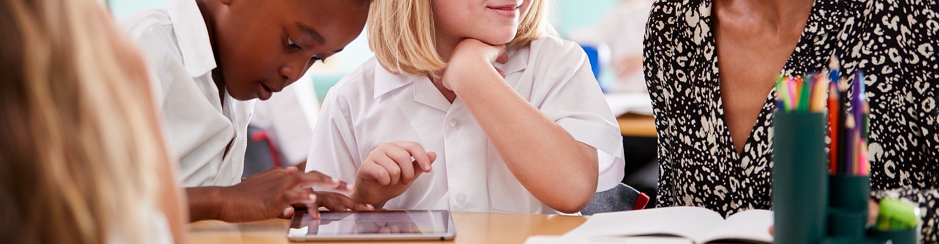 Female Teacher With Two Elementary School Pupils Wearing Uniform Using Digital Tablet At Desk