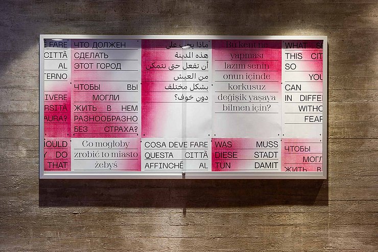 Typografisches Kunstwerk an einer Betonwand, bestehend aus mehrsprachigen Texttafeln in Rot-Weiß. Die Tafeln zeigen die Frage: „Was muss diese Stadt tun, damit du ohne Angst verschieden leben kannst?“ in mehreren Sprachen.