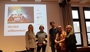 Dei Projketpartner, vier Frauen und ein Mann, stehen vor dem Titelbild der PPT für den Kick-Off des Projekts in einem Seminarraum. 