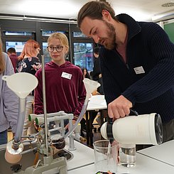 Student erklärt zwei Schülerinnen ein Experiment im Lehr-Lern-Labor der Physik